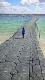 Side view of baby boy standing on pier by sea against cloudy sky during sunny day