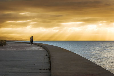 Man on footpath by sea against sky during sunset