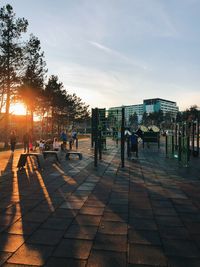 People on footpath in city against sky during sunset