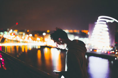 Young man using mobile phone while standing against illuminated buildings by river at night