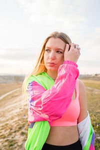 Contemporary blond haired young woman in pink bra with colorful jacket looking away with remote land on background