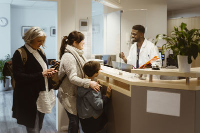 Patients standing and talking to male receptionist through transparent shield in clinic