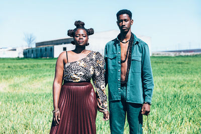 Young black girlfriend and boyfriend in stylish clothes with authentic african ornaments and accessories holding hands and looking at camera while standing in green grassy field in countryside together