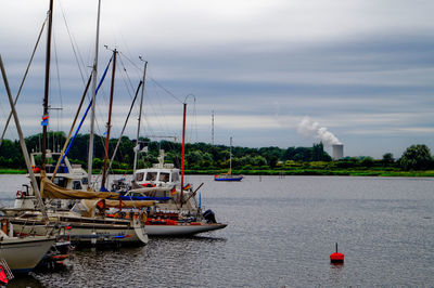 Boats moored on river against silo emitting smoke in sky