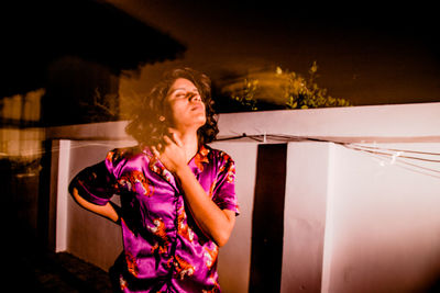 Young woman looking away while standing against illuminated wall at night