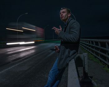 Side view of young man smoking while standing by railing at night