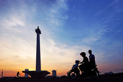 Silhouette people with bicycle against sky during sunset