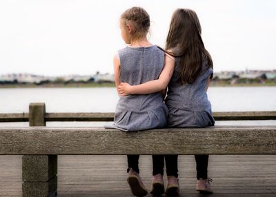 Full length rear view of girls sitting on pier against clear sky