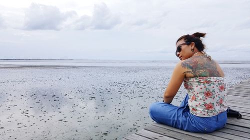 Tattooed woman sitting on pier at beach against sky