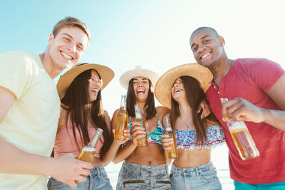 Low angle view portrait of friends holding drinks while standing at beach against sky
