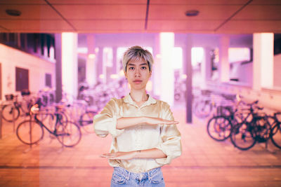 Portrait of young woman gesturing equal sign while standing on footpath against illuminated lights