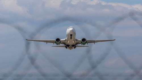 Low angle view of airplane flying against sky seen through barb wire