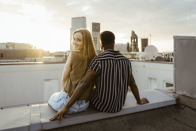 Portrait of smiling woman looking over shoulder while sitting with boyfriend on building terrace