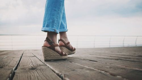 Low section of woman wearing sandals walking on pier against sky