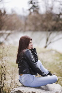 Side view of thoughtful teenage girl wearing leather jacket while sitting on rock