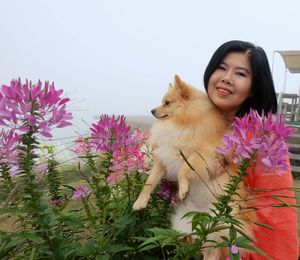 A woman smiles at the camera while holding a dog at the same time . grove of pink flowers.