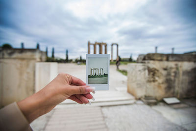 Cropped woman holding photograph at olympian zeus temple against cloudy sky