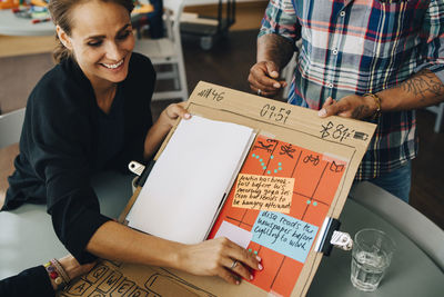 High angle view of smiling businesswoman showing strategy on placard to manager at table in creative office