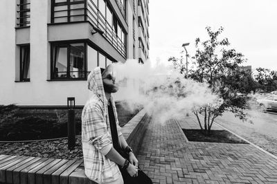 Young man smoking while sitting on retaining wall against building