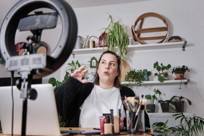 Mature female vlogger gesturing while recording make-up tutorial at home