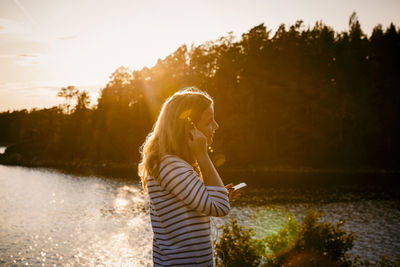 Side view of mid adult woman using mobile phone while standing against lake at sunset