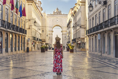 Rear view of woman wearing hat standing on footpath amidst buildings in city