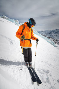 A professional skier athlete in a red jacket, helmet and mask stands on a slope against the backdrop