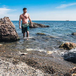 Full length of shirtless man on rocks at beach against sky