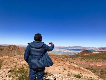 Rear view of man photographing while standing on mountain against clear blue sky