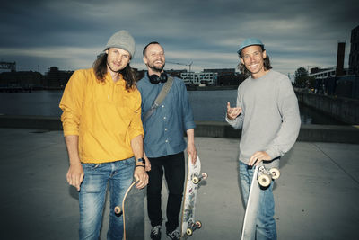 Portrait of smiling male friends with skateboards against canal at dusk