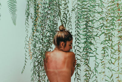 Rear view of shirtless woman standing against ivy wall
