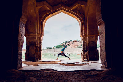 Side view of unrecognizable fit female in activewear performing warrior i pose while practicing yoga near old arched oriental palace in mountainous terrain