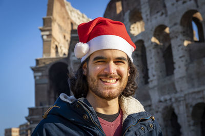 Smiling young man with a santa hat in front of the colosseum. christmas roman holiday concept.