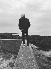 Rear view of man standing on retaining wall at beach against sky