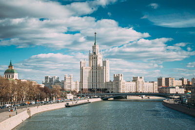 View of city buildings by river against cloudy sky