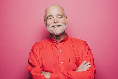 Portrait of smiling senior man with arms crossed against pink background