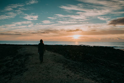 Rear view of woman walking towards sea against sky during sunset