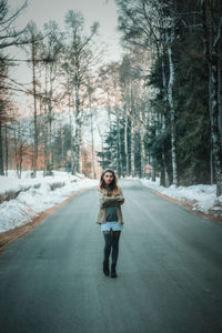 Portrait of young woman standing on road amidst trees during winter