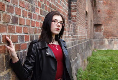 Young beautiful girl in black jacket and jeans stands near the old fortress wall on sunny day