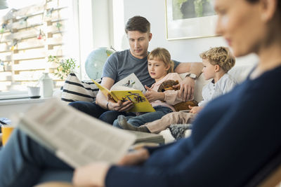 Father reading story book to kids with mother sitting in living room