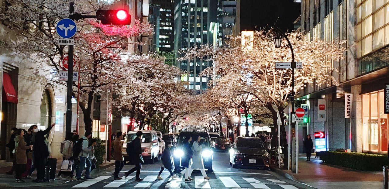 PEOPLE WALKING ON ILLUMINATED STREET AMIDST BUILDINGS IN CITY