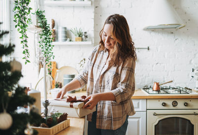 Attractive smiling woman with curly hair in plaid shirt with gift box near window at bright kitchen 