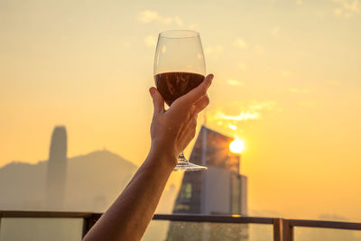 Cropped hand of woman holding wineglass against sky during sunset