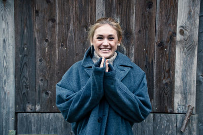 Portrait of smiling young woman wearing warm clothing standing against wooden wall