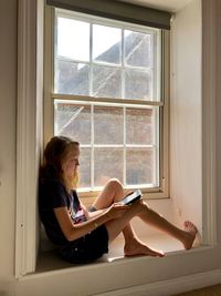 Side view of girl using phone while sitting on window at home