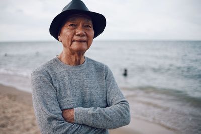 Portrait of smiling senior man with arms crossed standing at beach against sky during sunset