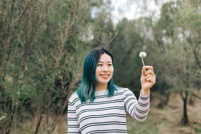 Smiling young woman holding flower while standing in forest