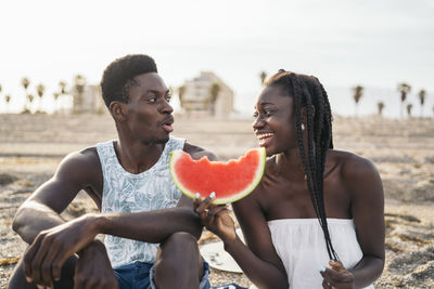 Teenage girl holding slice of watermelon while sitting by man at beach