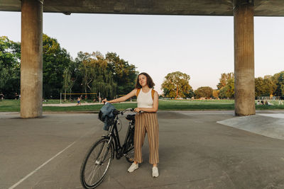 Teenage girl with bicycle standing in front of bridge at park