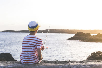 Rear view of boy fishing while sitting on stone wall by sea
